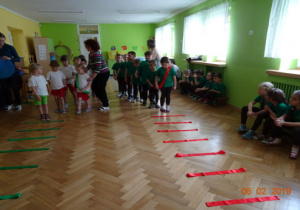 Dzieci stoją z nauczycielkami w rzędach. Na podłodze rozłożone są czerwone wstążki. Z boku na ławkach siedzą dzieci. wstążki